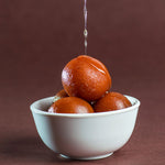 Gulab Jamun, un postre tradicional indio servido en un tazón blanco, con bolitas de masa frita empapadas en un jarabe dulce, con un chorro de jarabe que cae sobre ellas, presentado sobre un fondo marrón.