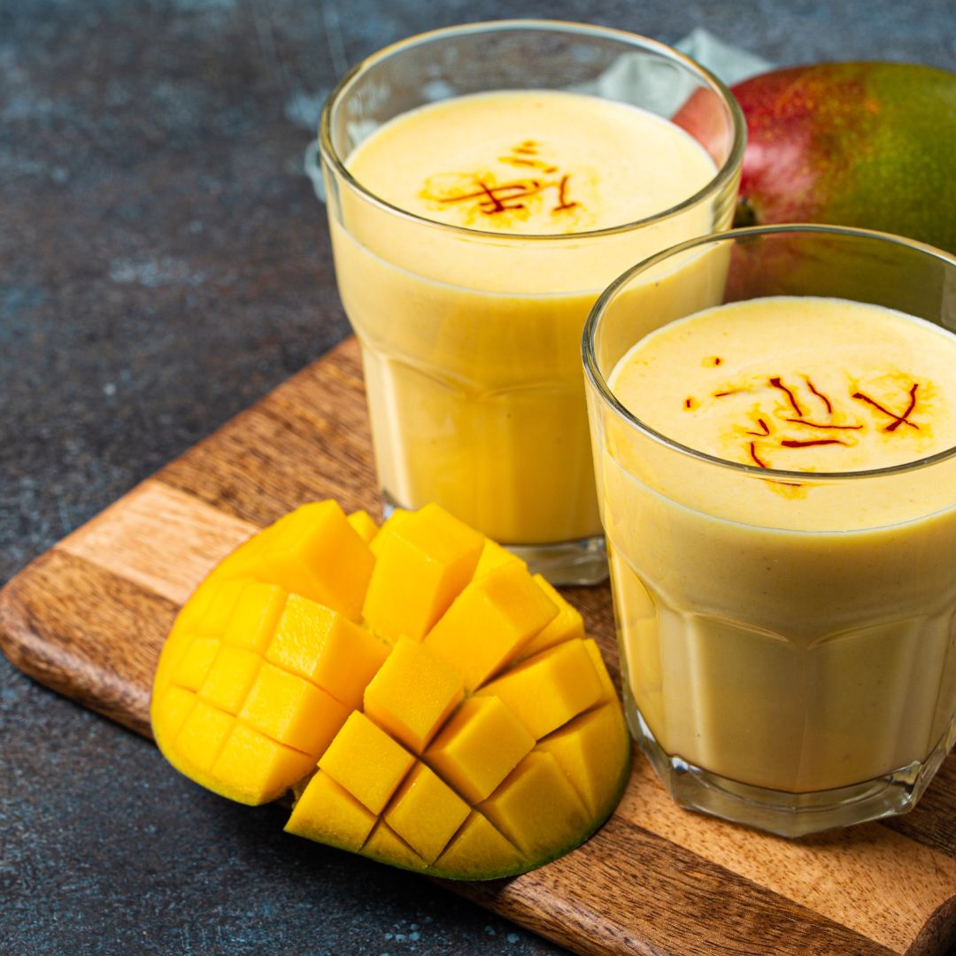 Mango Lassi, una bebida tradicional india servida en dos vasos, hecha de yogur y pulpa de mango, decorada con hebras de azafrán. Acompañada de un mango fresco cortado en cubos, todo presentado sobre una tabla de cortar de madera y un fondo oscuro.
