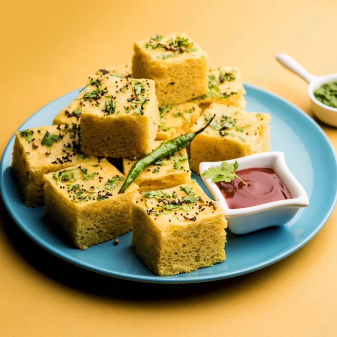 Khaman Dhokla, un delicioso aperitivo indio en forma de cubos esponjosos y amarillos, servido en un plato azul con guarniciones de cilantro y semillas de mostaza, acompañado de salsas en pequeños recipientes, presentado sobre un fondo amarillo.