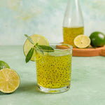 Bebida refrescante de limón con semillas de albahaca, servida en un vaso con una rodaja de limón y hojas de menta, con limones frescos y una botella de jal jeera en el fondo, presentada sobre una superficie verde.
