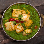 Palak Paneer, un plato clásico de la cocina india, servido en un recipiente metálico, con cubos de paneer dorado en una cremosa salsa de espinacas, adornado con una guindilla roja y hojas de cilantro fresco, presentado sobre una mesa de madera rústica.