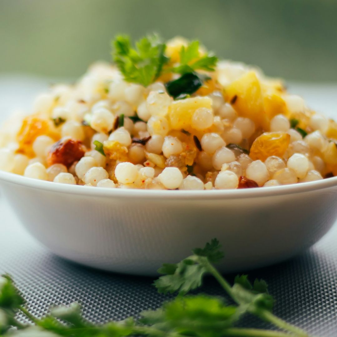 Sabudana Khichadi, un plato delicioso servido en un tazón blanco, con perlas de tapioca cocidas, hierbas frescas y trozos de vegetales, decorado con cilantro, presentado sobre una superficie gris.