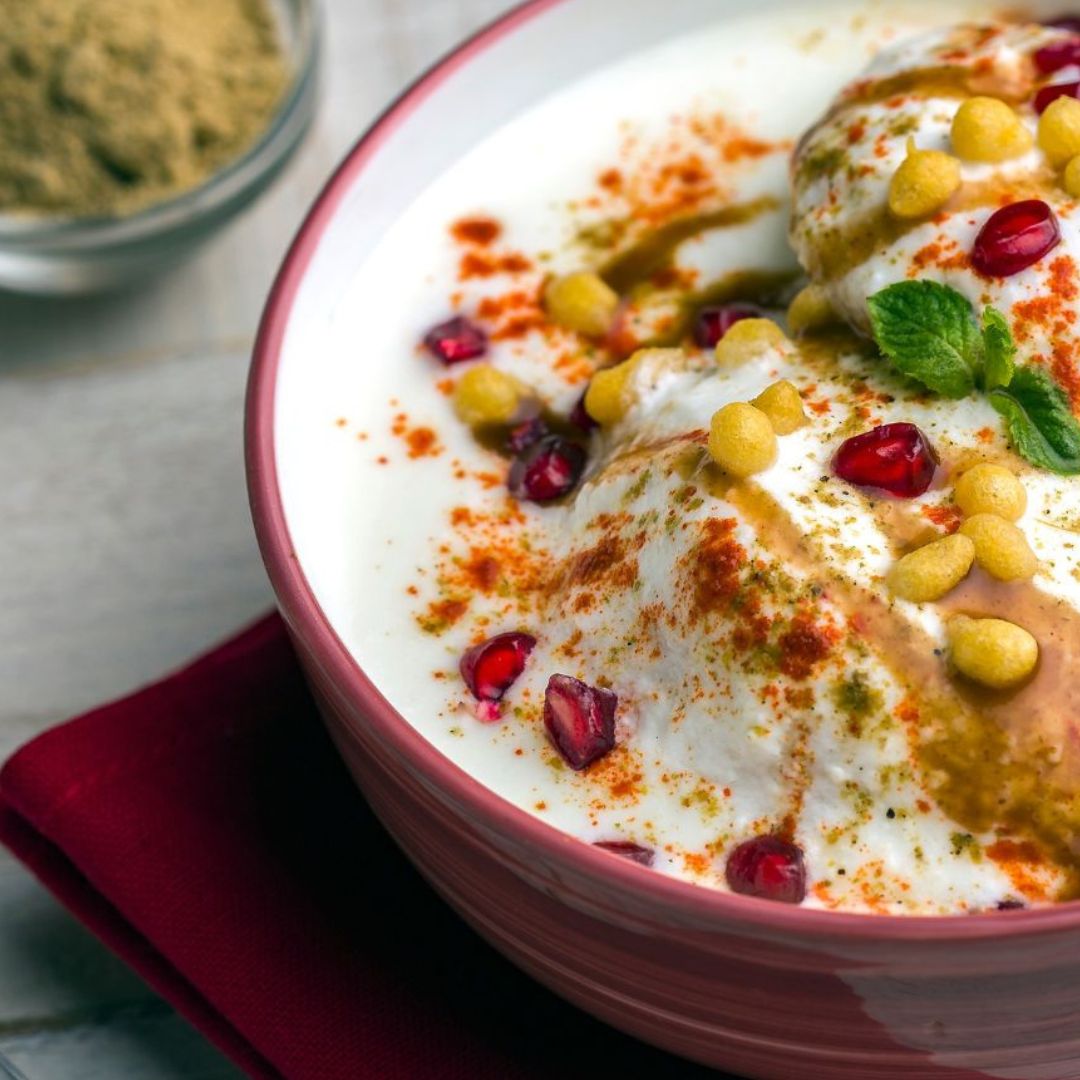 Dahi Vada, un aperitivo indio servido en un tazón rojo, con bolitas de masa frita cubiertas de yogur cremoso, granos de granada, especias y hierbas frescas, acompañado de un cuenco pequeño con polvo de especias, presentado sobre una servilleta roja.